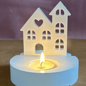 Puede incluir: Portavelas de cer&aacute;mica blanca en forma de casa con un coraz&oacute;n recortado y una peque&ntilde;a vela de t&eacute; dentro.