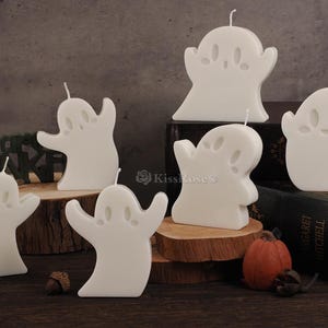 Op de afbeelding: Set witte kaarsen in de vorm van een spook. De kaarsen staan op een houten oppervlak met een boek op de achtergrond.