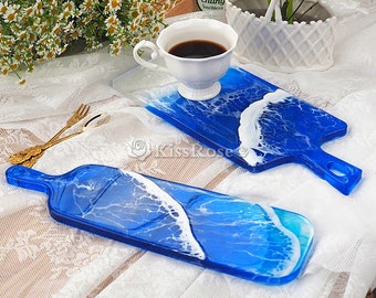 May include: Two blue resin serving boards with white wave designs. One board is rectangular with a handle, the other is a paddle shape. A white teacup filled with coffee sits on one board. Decorative and functional.
