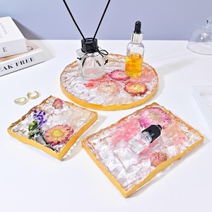 May include: Three clear resin trays with gold trim and dried flowers. The trays are different sizes and shapes. The largest tray has a diffuser bottle and a small bottle of essential oil.