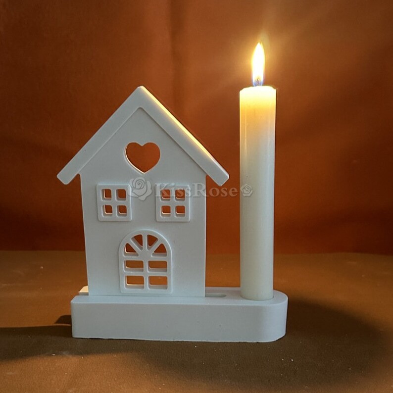 May include: White wooden house shaped candle holder with a heart cut-out and windows. A white pillar candle is in the holder.