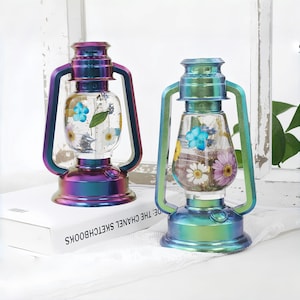 May include: Two iridescent lanterns with a rainbow metallic finish. The glass globes are filled with dried flowers in shades of blue, yellow, and purple. The lanterns have a handle and base, and are sitting on a white surface.