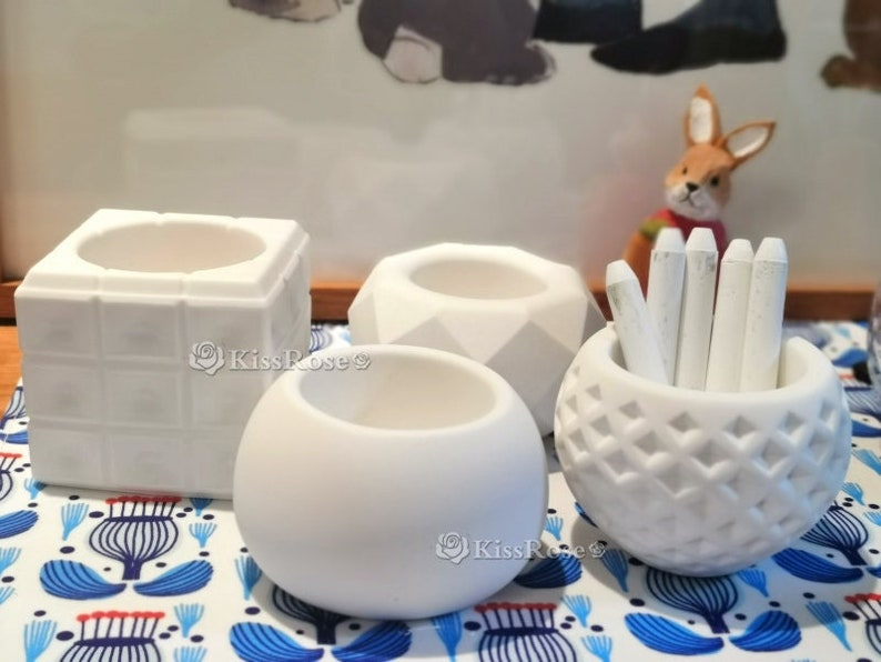 May include: Four white ceramic containers with different geometric designs. The container on the right has a pattern of small squares and holds white chalk sticks.