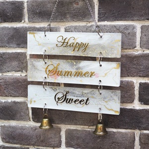 Puede incluir: Un letrero de madera blanco con letras doradas que dice "Happy Summer Sweet" colgado de una cadena con dos campanas doradas. El letrero tiene un diseño de mármol y detalles de brillo.