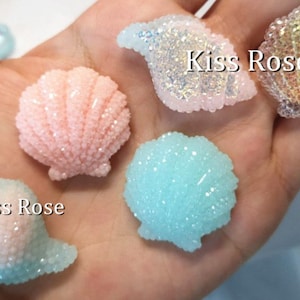 May include: Four sparkly seashell-shaped resin charms in pink, blue, white, and iridescent colours. The charms are labelled "Kiss Rose".