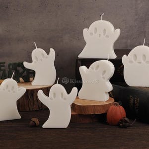 Op de afbeelding: Set van zes witte kaarsen in de vorm van een spook. De kaarsen zijn gerangschikt op een houten oppervlak met een boek op de achtergrond.