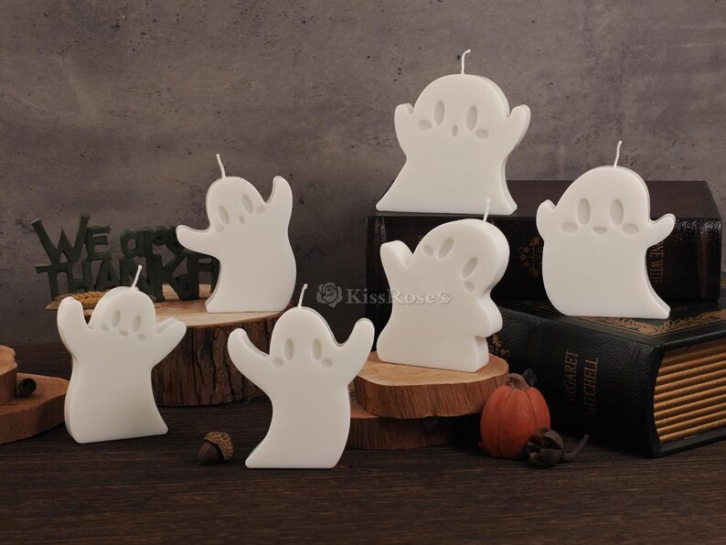 Op de afbeelding: Set van zes witte kaarsen in de vorm van een spook. De kaarsen zijn gerangschikt op een houten oppervlak met een boek op de achtergrond.