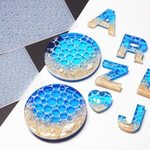 Könnte beinhalten: Ein Set aus Harzformen zum Basteln mit Strandthema. Die Formen sind klar und flexibel, mit einer strukturierten Oberfläche, die an Meereswellen erinnert. Das Set enthält Formen für Buchstaben, ein Herz und zwei runde Formen.