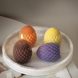 May include: Four dragon egg-shaped candles on a white circular tray. The candles are in shades of orange, yellow, purple, and brown.