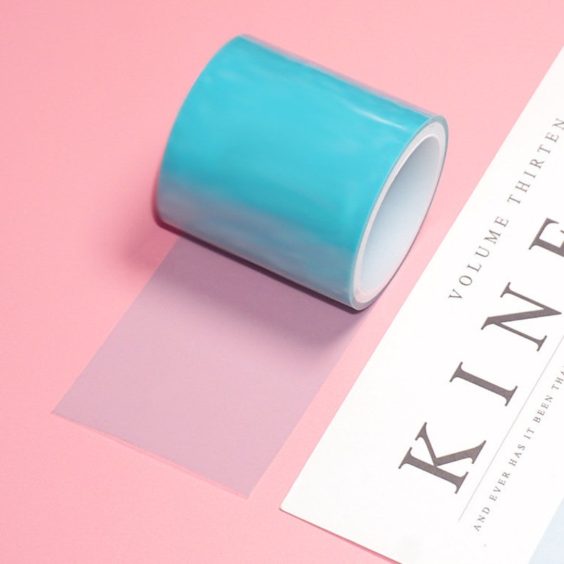 Pode incluir: Um rolo de fita adesiva azul transparente sobre uma superf&iacute;cie rosa. A fita est&aacute; parcialmente desenrolada e a ponta da fita &eacute; vis&iacute;vel. A fita est&aacute; sobre uma folha de papel branca com o texto "VOLUME THIRTEEN" e "KIN" impresso nela.