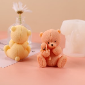 Puede incluir: Dos figuritas de ositos de peluche, una sosteniendo un osito de peluche más pequeño. El osito más grande es de color melocotón y el osito más pequeño es marrón claro. Ambos ositos tienen ojos y narices negros. Los ositos están sentados sobre una superficie rosa.