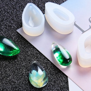 May include: Clear resin teardrop pendants in green, blue, and yellow hues, alongside white silicone molds. The pendants have a glossy finish, and the molds are in a teardrop shape. The background is a dark, textured surface.