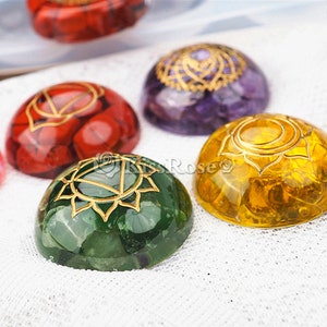 May include: Seven chakra stones, each with a different color and a gold symbol. The stones are arranged in a row on a white fabric background.