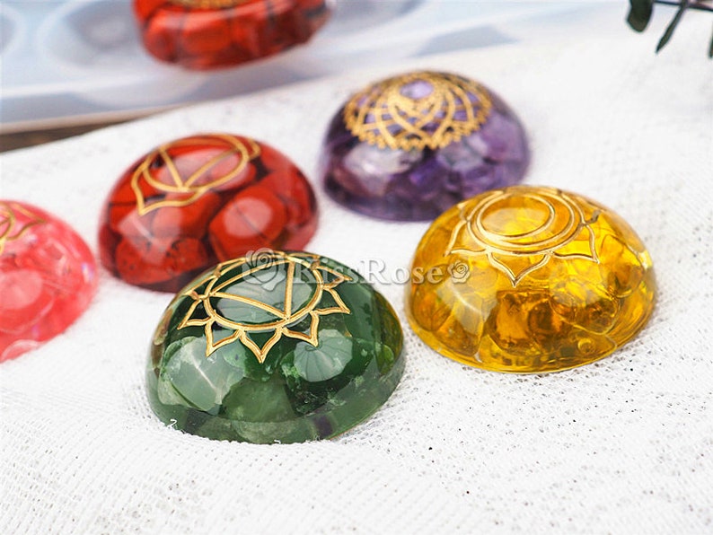 May include: Seven chakra stones, each with a different color and a gold symbol. The stones are arranged in a row on a white fabric background.