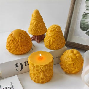 May include: A collection of yellow beeswax candles in various shapes, including a lit cylindrical candle, a beehive, and a tree-shaped candle. The candles are decorated with bee and honeycomb patterns.