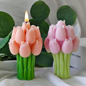 May include: Two tulip-shaped candles. One is lit, with a flame at the top. The tulips are a soft pink color, and the stems are green. The other candle is a similar design, but unlit. The background includes green leaves.