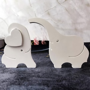 Puede incluir: Dos figuritas de elefante de madera blanca, una con un corazón recortado en el pecho, la otra con la trompa levantada. Las figuritas están sobre una superficie oscura.