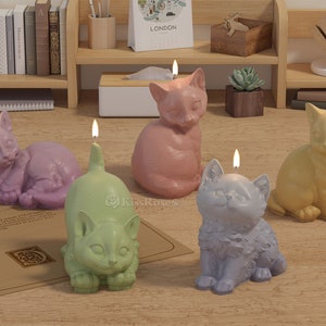 Puede incluir: Cinco velas con forma de gato en diferentes colores, incluyendo rosa, morado, verde, azul y amarillo. Las velas están todas sobre una mesa de madera.
