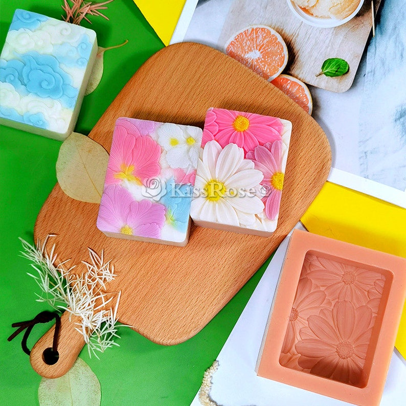 May include: Three handmade soap bars with floral designs. The soaps are on a wooden cutting board. The soap bars are in a floral design with pink, white, and blue flowers. The soap bars are in a silicone mold with a floral design.