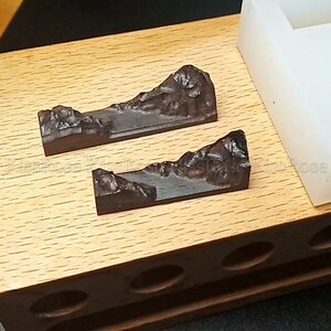 May include: Two dark brown wooden mountain molds in a wooden box with a white silicone mold in the background.