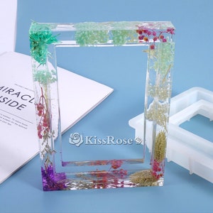 Pode incluir: Um porta-retratos de resina transparente com flores secas e vegetação incorporadas no interior. O porta-retratos é retangular e tem um fundo branco. O porta-retratos está sobre uma superfície branca. O texto "KissRose" é visível na imagem.