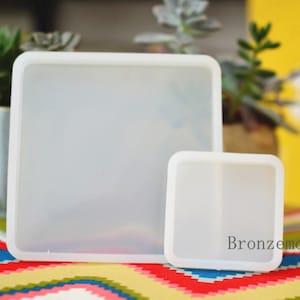 Peut inclure: Deux moules en silicone blanc pour la coulée de résine. Le moule le plus grand est carré et le moule le plus petit est également carré mais légèrement plus petit. Les moules sont sur une surface colorée.