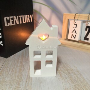 Pu&ograve; includere: Portacandele in ceramica bianca a forma di casa con un ritaglio a forma di cuore nella parte anteriore. Una piccola candela da t&egrave; sta bruciando all'interno del portacandele, proiettando un bagliore caldo.