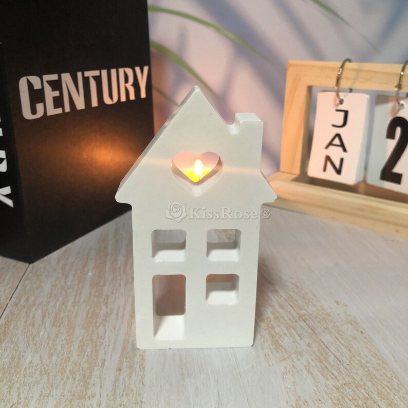 Pu&ograve; includere: Portacandele in ceramica bianca a forma di casa con un ritaglio a forma di cuore nella parte anteriore. Una piccola candela da t&egrave; sta bruciando all'interno del portacandele, proiettando un bagliore caldo.