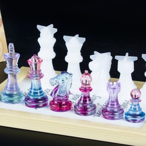 K&ouml;nnte beinhalten: Ein Set aus bunten gl&auml;sernen Schachfiguren mit einem Farbverlauf von Rosa, Blau und Lila. Die Figuren sind auf einem Holzbrett mit wei&szlig;en Feldern angeordnet. Die Figuren umfassen K&ouml;nig, Dame, L&auml;ufer, Springer, Turm und Bauer.