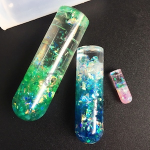 Op de afbeelding: Drie transparante glazen buizen gevuld met groene, blauwe en roze iriserende glitter. De buizen zijn van verschillende grootte en zijn gerangschikt in een rij van groot naar klein.