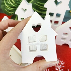 May include: White wooden house ornament with a heart cutout and four windows. The ornament is held in a hand.