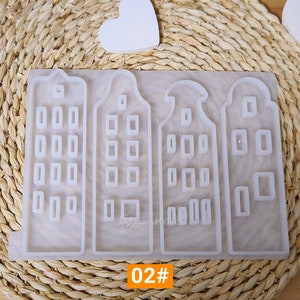 May include: A silicone mold with four separate molds of Dutch houses. Each house has a pointed roof and multiple windows. The mold is white and is sitting on a woven straw mat. The text "02#" is visible at the bottom of the image.