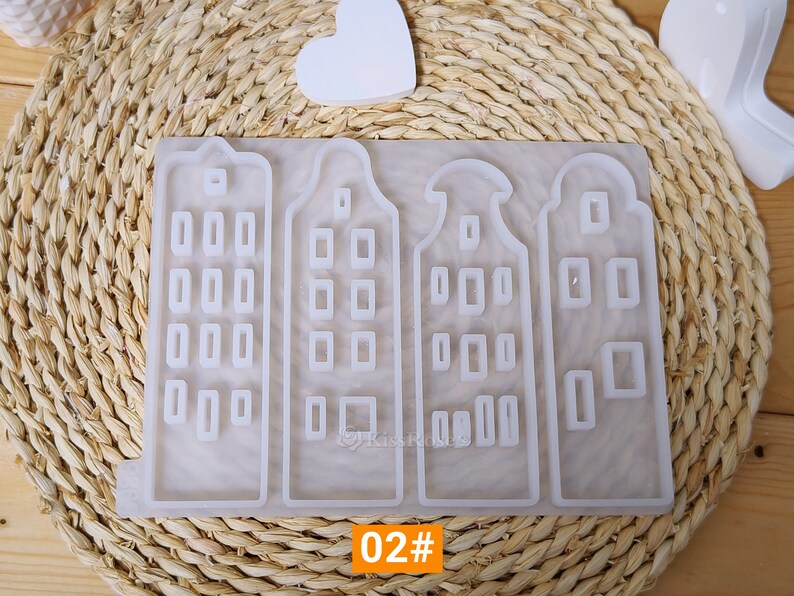 May include: A silicone mold with four separate molds of Dutch houses. Each house has a pointed roof and multiple windows. The mold is white and is sitting on a woven straw mat. The text "02#" is visible at the bottom of the image.