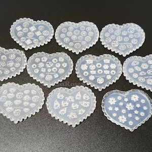 Puede incluir: Diez moldes de silicona transparentes en forma de corazón con intrincados diseños florales. Cada molde tiene un patrón diferente de pequeñas flores y remolinos.