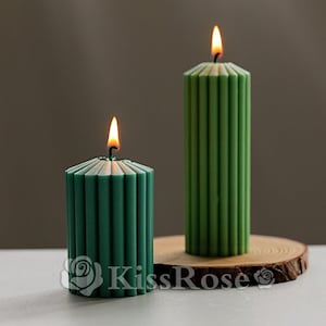 May include: Two green pillar candles with a ribbed design. The candles are lit and are on a wooden base.