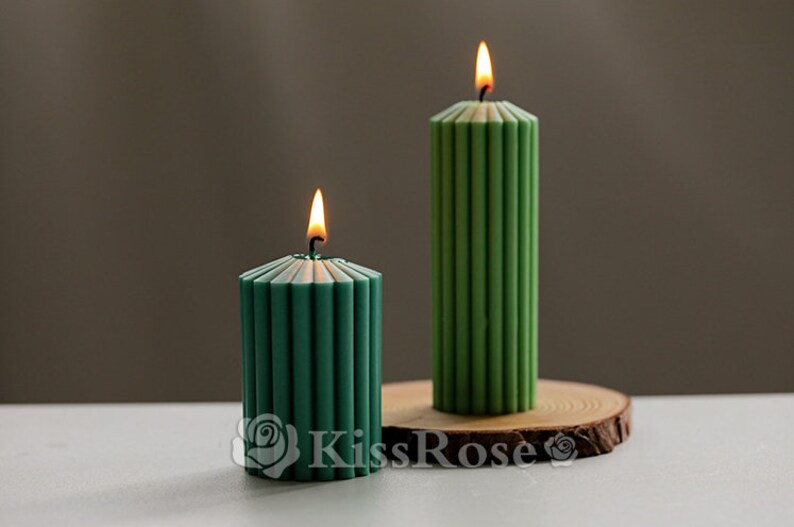 May include: Two green pillar candles with a ribbed design. The candles are lit and are on a wooden base.