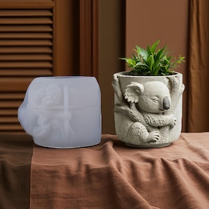 Puede incluir: Una maceta gris con forma de koala con una pequeña planta verde en su interior. La maceta tiene un relieve detallado de un koala abrazando una rama de árbol. Al lado, un molde de silicona transparente de la maceta.