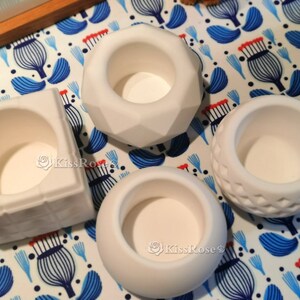 May include: Four white ceramic candle holders with different shapes and designs. The holders are on a white and blue patterned surface.