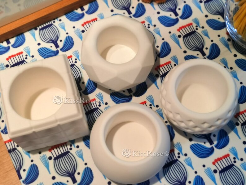 May include: Four white ceramic candle holders with different shapes and designs. The holders are on a white and blue patterned surface.