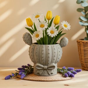Op de afbeelding: Een grijze, cactusvormige keramische plantenbak met een strikdetail, gevuld met gele tulpen en witte madeliefjes. De plantenbak staat op een geweven mat, met takjes paarse lavendel rond de basis.
