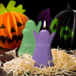 May include: Purple ghost shaped candle with a lit flame, surrounded by straw-like material. The candle is in front of a green ghost, an orange pumpkin, and a black bat.