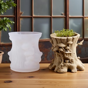 Könnte beinhalten: Zwei dekorative Pflanzgefäße auf einer Holzoberfläche. Eines ist eine Vase aus mattiertem Glas mit strukturiertem Design. Das andere ist ein Baumstumpf-Pflanzgefäß mit Pilz- und Vogeldetails, gefüllt mit Grün. Der Hintergrund zeigt ein Fenster.