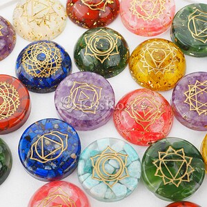 May include: Set of seven chakra stones, each with a different color and a gold symbol. The stones are arranged in a circle on a white surface.