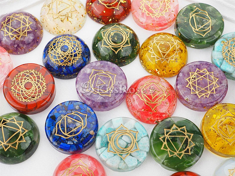 May include: Set of seven chakra stones, each with a different color and a gold symbol. The stones are arranged in a circle on a white surface.