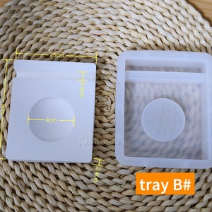 May include: Two white silicone molds for resin casting. The mold on the left is 7.5 cm wide, 9 cm tall, and 10.8 cm deep. The mold on the right is 24 cm tall. The mold on the left has a circular indentation that is 4 cm in diameter. The mold on the right has a rectangular indentation and a circular indentation. The text "tray B#" is printed on the bottom right corner of the image.