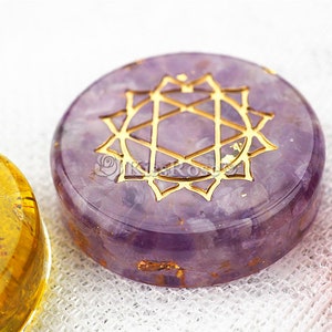 May include: A purple amethyst crystal with a gold geometric design, known as a Flower of Life symbol. The crystal is round and has a smooth, polished surface.