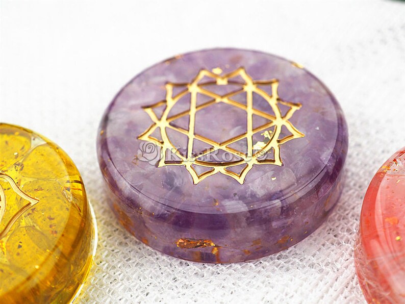 May include: A purple amethyst crystal with a gold geometric design, known as a Flower of Life symbol. The crystal is round and has a smooth, polished surface.