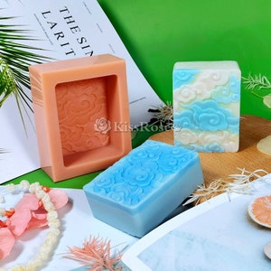 May include: Two silicone soap molds with a cloud design. One mold is pink and the other is clear. Two bars of soap are shown, one is blue and the other is white with blue clouds.