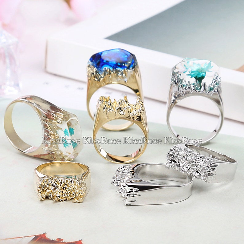 May include: A set of six gold and silver rings with different designs. Some rings feature a clear resin with embedded flowers or gemstones. Others have a textured, rough surface.