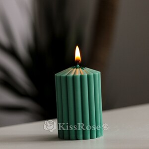 May include: A teal green pillar candle with a fluted design is lit and burning. The candle is on a white surface.